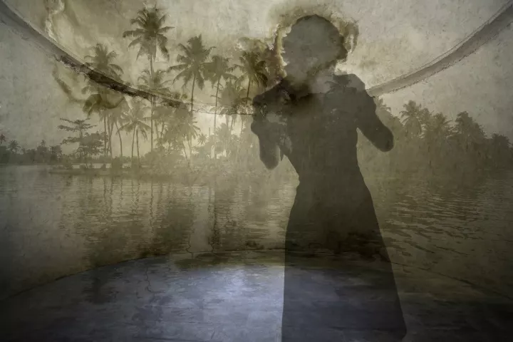 En transparence dans une pièce ronde, une silhouette féminine de dos fait face au paysage d'une île avec des palmiers.