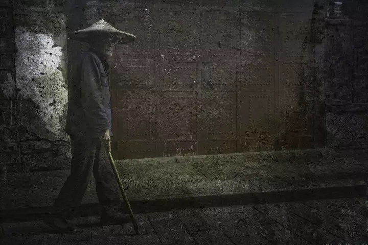 Un vieux chinois en tenue traditionnelle marche dans la rue et passe devant une porte rouge.